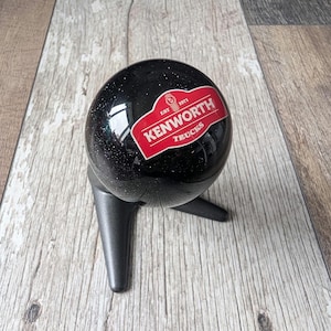 May include: A black, spherical gear shift knob with a red and white "Kenworth Trucks" logo. The knob is mounted on a black base with three legs. The logo includes the text "EST 1971".