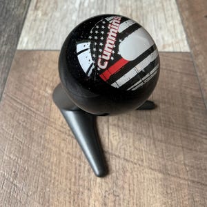 Peut inclure: Un pommeau de levier de vitesses noir avec un motif du drapeau américain, une bande rouge et le mot "Cummins" en blanc. Le pommeau est monté sur une base noire.