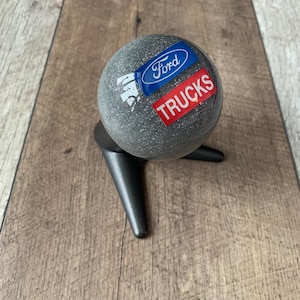 Peut inclure: Un pommeau de levier de vitesses sphérique gris avec le logo Ford bleu et le mot "TRUCKS" en rouge. Monté sur une base noire à trois branches, sur un fond imitation bois.