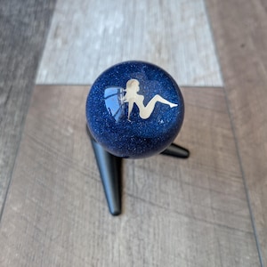 Peut inclure: Pommeau de levier de vitesses bleu foncé et scintillant avec une silhouette blanche de femme. Le pommeau est monté sur une base noire. Le fond est une surface en bois.