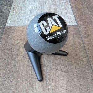 Peut inclure: Une sphère grise et scintillante avec le logo CAT Diesel Power en noir, blanc et jaune. La sphère est posée sur un support noir à trois pieds. L'arrière-plan est une surface en bois clair.