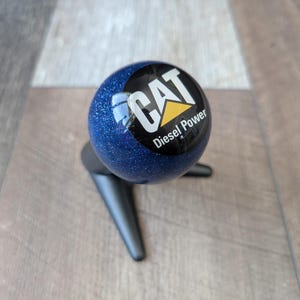 Peut inclure: Une sphère bleu pailleté avec le logo CAT et le texte "Diesel Power". La sphère est montée sur un support noir à trois pieds. Le fond est une surface en bois brun clair.