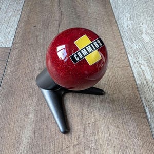May include: A spherical red gear shift knob with a glitter finish, featuring a black and yellow rectangular logo with the text CUMMINS printed in white. The knob is mounted on a matte black, three-pronged metal stand for display purposes.