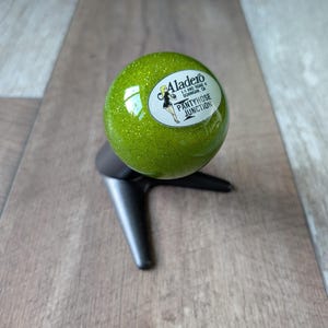 May include: A spherical green glitter gear shift knob mounted on a black tripod stand. The knob features a white oval decal with a graphic of a person and the text Aladero I-5 AND ROAD 8 DUNNIGAN, CA PANTYHOSE JUNCTION printed in black ink.