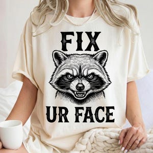Peut inclure: Un t-shirt couleur crème avec une illustration graphique noire représentant une tête de raton laveur aux dents découvertes. Le texte FIX est imprimé au-dessus du raton laveur et le texte UR FACE est imprimé en dessous en police noire.