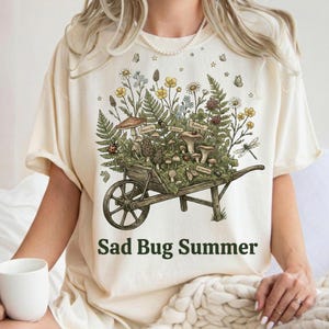 Peut inclure: Un t-shirt couleur crème arborant une illustration graphique d'une brouette en bois remplie de divers champignons, fougères, fleurs sauvages et insectes. Le texte "Sad Bug Summer" est imprimé en lettres vert foncé sous la brouette.