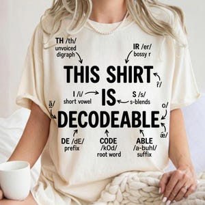Peut inclure: Un t-shirt couleur crème avec du texte noir indiquant THIS SHIRT IS DECODEABLE. Le motif comprend divers symboles phonétiques linguistiques, des flèches et des étiquettes telles que unvoiced digraph, bossy r, short vowel, s-blends, prefix, root word et suffix.