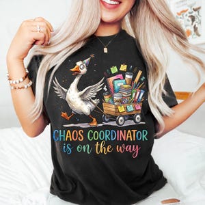 Peut inclure: Un t-shirt noir avec une oie de dessin animé portant un chapeau de fête et tirant un chariot en bois rempli de cahiers, de stylos et de tasses à café colorés. Le texte indique "CHAOS COORDINATOR is on the way" en lettres multicolores.
