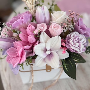 May include: A floral arrangement featuring a variety of flowers in shades of pink, purple, and white. The flowers are arranged in a white box with a gold chain handle. The arrangement includes tulips, roses, and other decorative flowers.