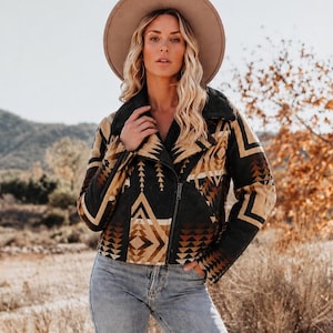 May include: A wide-brimmed tan felt hat and a cropped jacket featuring a dark charcoal, tan, and rust-colored geometric pattern. The jacket has a black faux-fur collar, long sleeves, and a diagonal front zipper closure, paired with light blue denim jeans.