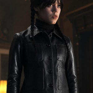 May include: A black leather jacket featuring a pointed collar, long sleeves, and four front patch pockets with button closures. The jacket is worn over a black turtleneck sweater and a black and white checkered shirt visible at the bottom hem.