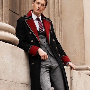 Op de afbeelding: Een zwarte lange jas met rode revers en manchetten, gedragen over een grijs vest en een grijze broek met ruitpatroon. De outfit bevat een wit overhemd met kraag en een donkerrode das met patroon. De jas heeft dubbele gouden knopen.