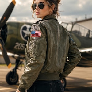May include: An olive green bomber jacket featuring a United States flag patch on the right shoulder, a zippered utility pocket on the sleeve, and ribbed cuffs. The jacket is worn with black jeans and dark-tinted aviator sunglasses.