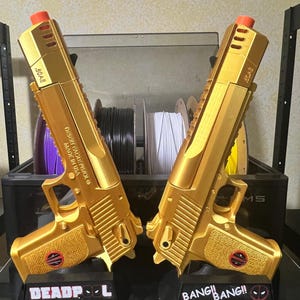 Puede incluir: Dos pistolas de juguete doradas con puntas naranjas, etiquetadas como "Desert Eagle Pistol" y ".50AE". Cada pistola descansa sobre una base negra con texto "DEADPOOL" y "BANG! BANG!!". El fondo incluye bobinas de filamento.