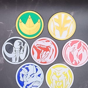 May include: Seven round, colorful coasters with different dinosaur and animal designs. The coasters are made of a 3D printed material and feature a white outline around each design.
