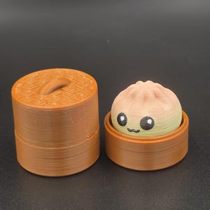 May include: A 3D printed brown cylindrical steamer basket with a lid and a small, round, light-colored dumpling character inside. The dumpling has a pleated top, two large black eyes with white highlights, and a small black curved mouth.