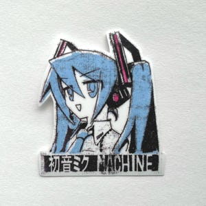 May include: A black and white sticker with a blue-haired anime character wearing headphones. The character has large eyes and a surprised expression. The text "初音ミク MACHINE" is printed below the character.