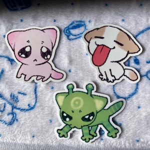 May include: Three cartoon animal stickers. A pink cat with big eyes, a white dog with brown spots sticking its tongue out, and a green alien-like creature with a circle on its forehead.