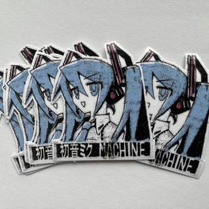 Pu&ograve; includere: Cinque adesivi con un personaggio anime dai capelli blu con un design in bianco e nero. Gli adesivi hanno il testo "初音ミク MACHINE".