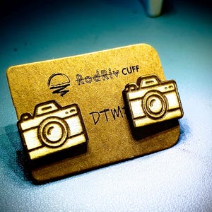 Könnte beinhalten: Ein Paar Holzohrringe in Form einer Kamera auf goldenem Hintergrund. Die Ohrringe sind mit "RodRiv Cuff" und "DTW" beschriftet.