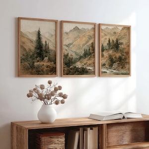 Op de afbeelding: Drie ingelijste landschapsprints met berglandschappen in gedempte bruin-, beige- en groentinten. Het kunstwerk toont bomen, bergen en een rivier. De lijsten zijn van lichtbruin hout. Het kunstwerk wordt boven een houten kast getoond.