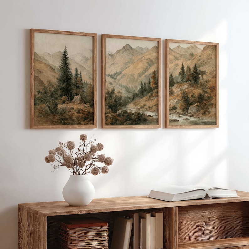 Op de afbeelding: Drie ingelijste landschapsprints met berglandschappen in gedempte bruin-, beige- en groentinten. Het kunstwerk toont bomen, bergen en een rivier. De lijsten zijn van lichtbruin hout. Het kunstwerk wordt boven een houten kast getoond.