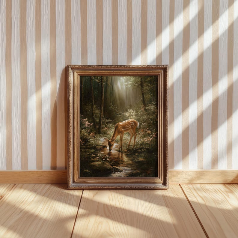 May include: A framed artwork depicts a deer drinking from a stream in a forest. The painting features a brown and gold frame, with a detailed landscape of trees, water, and a deer with white spots. The artwork is displayed on a wooden floor against a striped wall.
