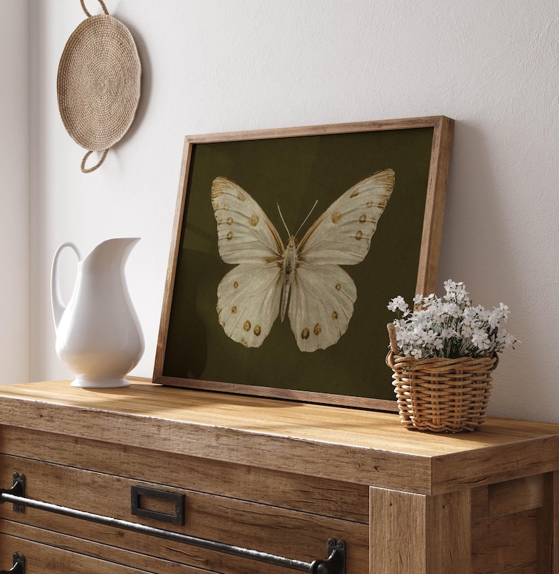 May include: A framed print of a white butterfly with gold markings on a dark green background. The print is in a wooden frame and is sitting on a wooden table.