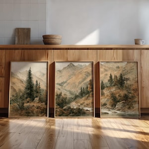 Op de afbeelding: Drie ingelijste landschapskunstprints. Elke print toont een aquarel schilderij van een berglandschap met bomen en een rivier. Het kleurenpalet omvat tinten bruin, groen en grijs, met een vintage esthetiek. De frames zijn lichtbruin.