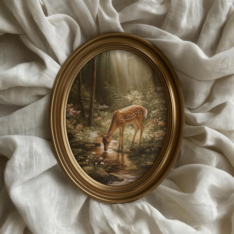 May include: An oval, gold-framed painting depicts a deer drinking from a stream in a forest. The deer is brown with white spots. The painting is set against a white, textured fabric.
