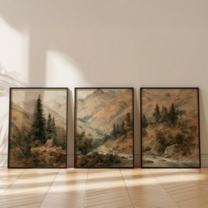 Op de afbeelding: Set van drie ingelijste landschapskunstprints. Elke print toont een aquarel schilderij van een berglandschap met bomen en een rivier. Het kleurenpalet omvat tinten bruin, groen en beige.