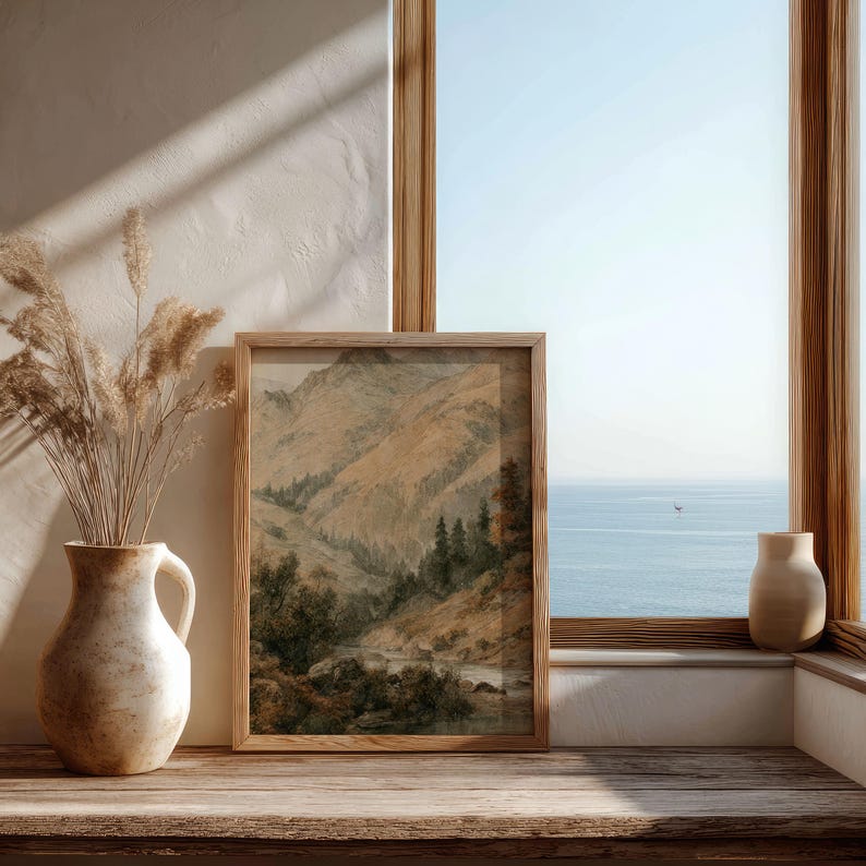 Op de afbeelding: Een ingelijste kunstdruk met een berglandschap met een rivier en bomen. Het kunstwerk staat in een houten lijst en staat op een houten oppervlak naast een vaas met gedroogde planten. Op de achtergrond is een raam met uitzicht op de oceaan.