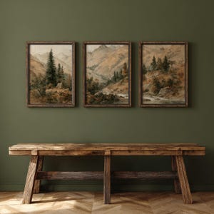 Op de afbeelding: Drie ingelijste landschapsschilderijen met berglandschappen met bomen en een rivier. Het kunstwerk gebruikt een gedempte kleurenpalet van bruin, groen en beige. Onder het kunstwerk staat een rustieke houten bank tegen een donkergroene muur.