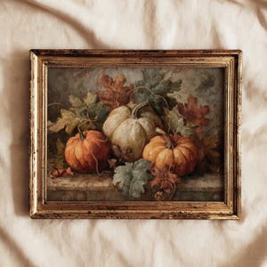 Könnte beinhalten: Gerahmtes Stillleben-Gemälde mit drei Kürbissen in Orange- und Cremetönen, umgeben von Herbstblättern. Das Kunstwerk ist vor einem gedämpften Hintergrund platziert und in einem goldfarbenen, verzierten Rahmen eingefasst. Das Gemälde erinnert an das Erntethema.
