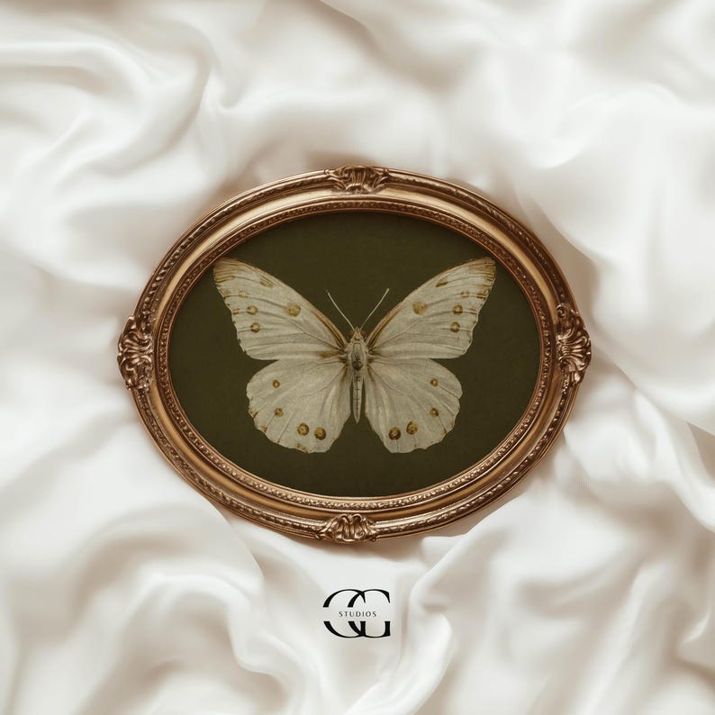 May include: A white butterfly with gold markings is framed in a gold oval frame with a dark green background. The frame is on a white satin fabric.