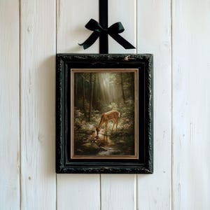 May include: A framed painting of a deer drinking from a stream in a forest, with a black frame and a black ribbon. The painting has a vintage aesthetic, with a focus on light and shadow. The artwork is hung against a white wooden background.