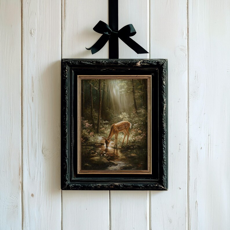 May include: A framed painting of a deer drinking from a stream in a forest, with a black frame and a black ribbon. The painting has a vintage aesthetic, with a focus on light and shadow. The artwork is hung against a white wooden background.