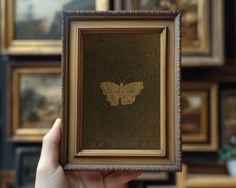 May include: A framed artwork featuring a gold butterfly design on a dark green background. The butterfly is centered within a circular pattern. The frame is a detailed, ornate gold color. The artwork is held in a hand.