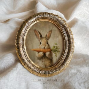 May include: A framed print featuring a brown rabbit holding a carrot. The artwork is set within a gold-colored, oval-shaped frame with a decorative edge. The text "GOLDEN GARLAND studios" is at the bottom.