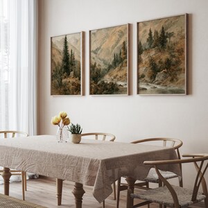 Op de afbeelding: Drie ingelijste landschapskunstafdrukken met berglandschappen met bomen en een rivier. Het kunstwerk heeft een gedempt kleurenpalet van bruin, groen en beige. De prints worden tentoongesteld boven een eettafel met een beige tafelkleed en houten stoelen.
