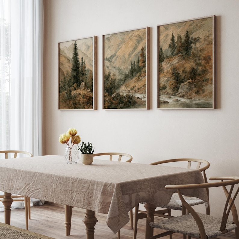 Op de afbeelding: Drie ingelijste landschapskunstafdrukken met berglandschappen met bomen en een rivier. Het kunstwerk heeft een gedempt kleurenpalet van bruin, groen en beige. De prints worden tentoongesteld boven een eettafel met een beige tafelkleed en houten stoelen.