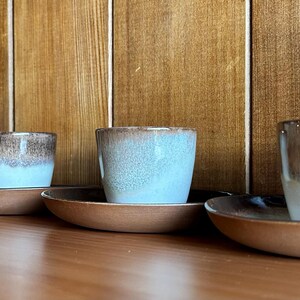 May include: Four ceramic espresso cups with brown and blue glaze, each sitting on a matching saucer. The cups and saucers are arranged in a row on a wooden surface.