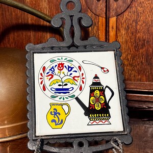 May include: A decorative black metal trivet with a square ceramic tile insert. The tile features a colorful design of a plate, a teapot, a spoon, and a pitcher. The trivet has a decorative handle and a scrolled base.