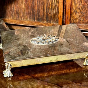 May include: A rectangular metal box with a decorative silver-colored medallion on the lid. The box has ornate feet and a tarnished finish, suggesting an antique or vintage item. The box is displayed on a wooden surface.
