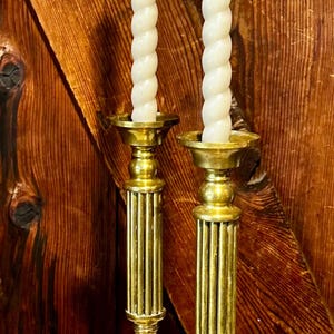 May include: Two antique brass candlesticks with white spiral candles. The candlesticks have a fluted column design and a round base. The background is a wooden door.