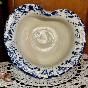 以下が含まれることがあります： 青と白の斑点模様の釉薬が施された白い陶器のボウル。ボウルはハート型で、わずかに波打った縁をしています。