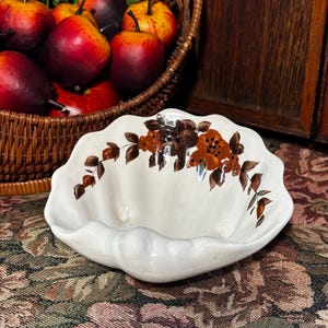 Könnte beinhalten: Eine weiße Keramikschale mit gewelltem Rand und einem Blumendesign in Braun- und Orangetönen. Die Schale ist leer und steht auf einer gemusterten Tischdecke. Ein Weidenkorb mit künstlichen Äpfeln befindet sich im Hintergrund.