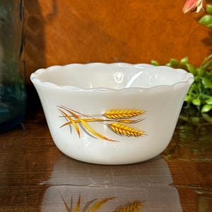 May include: A white, scalloped-edge bowl with a wheat stalk design in shades of gold and brown. The bowl is made of a glossy material and sits on a reflective wooden surface. The bowl is likely vintage or retro.