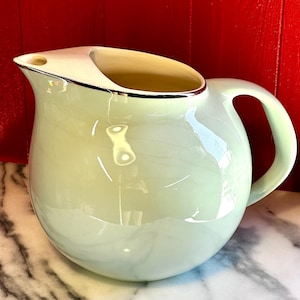 May include: A pale green ceramic jug with a rounded body and a curved handle. The jug has a wide opening with a silver trim and a spout. The interior is a light yellow colour. The jug is likely vintage and suitable for serving beverages.
