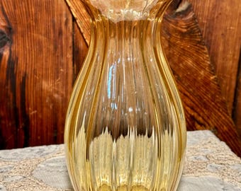 May include: A vintage amber glass vase with a ribbed design. The vase has a flared top and a rounded base. The glass is a warm, golden color, and the vase is sitting on a white lace tablecloth.